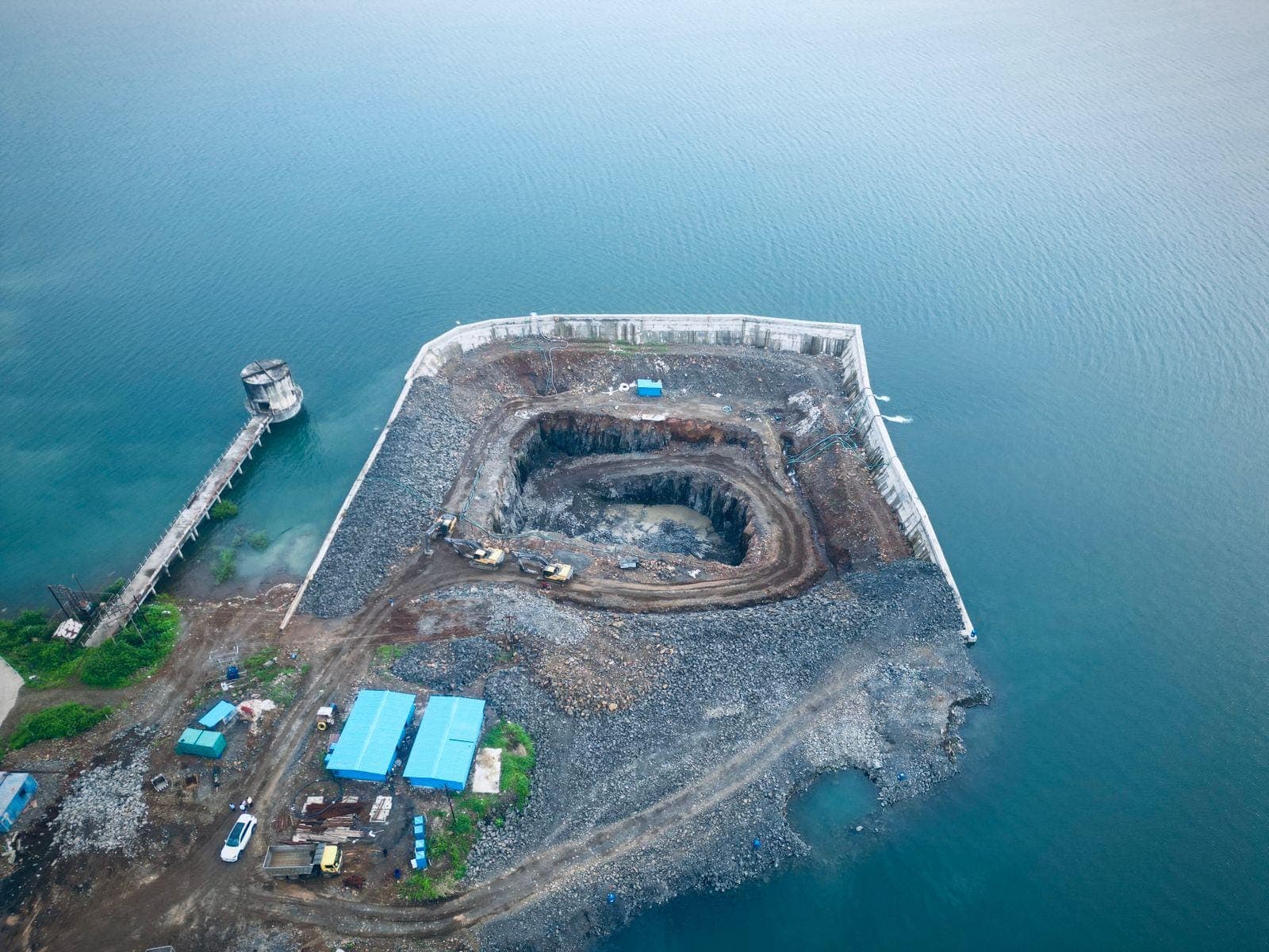 High aerial of the Bhama Askhed intake works — the diaphragm-walled excavation pit, cofferdam embankment, site offices, and approach jetty extending into the dam reservoir