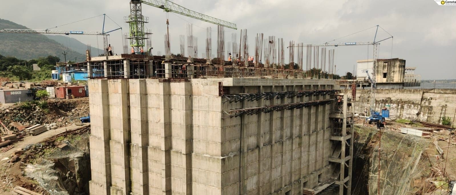 Tower cranes lifting over the reinforced-concrete pump house substructure at the Bhama Askhed head-works, with the dam reservoir and hills behind