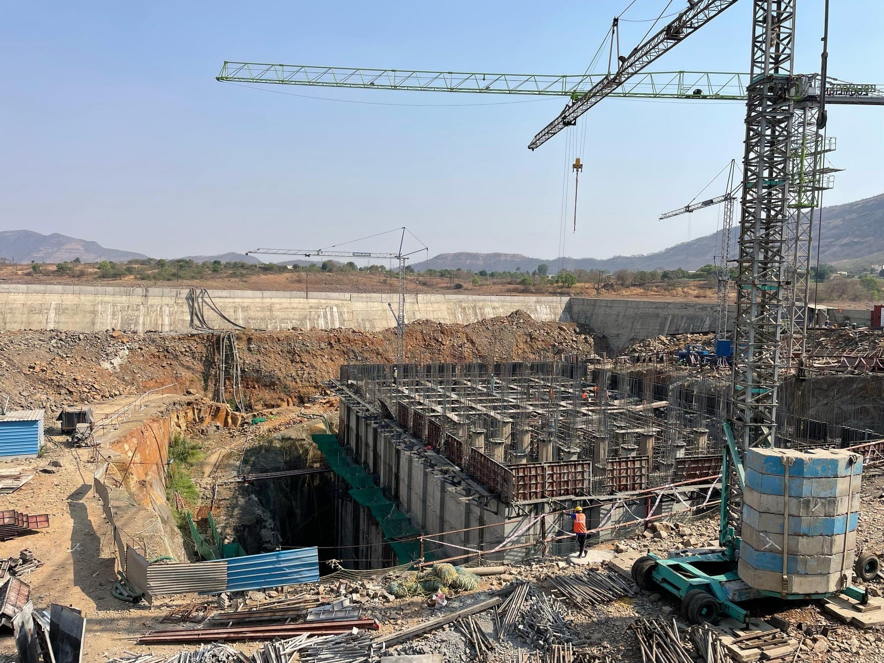 Tower crane over the column-grid substructure of the Bhama Askhed pump house, set within the rock excavation against the dam reservoir