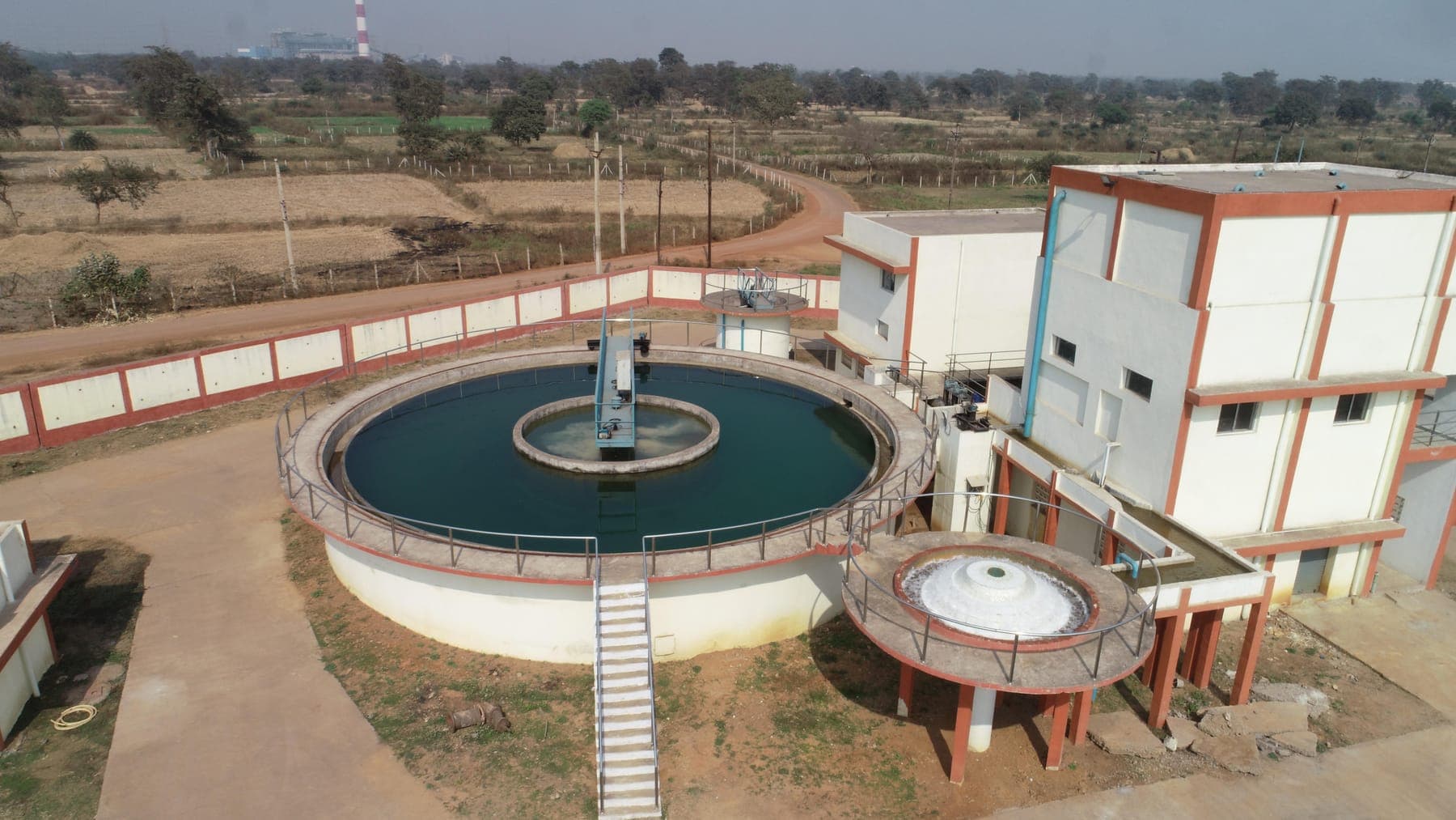 Circular clarifier with central feed column and a thickener tank next to the multi-storey treatment building at SAIL Bhilai, steel-plant chimneys on the horizon